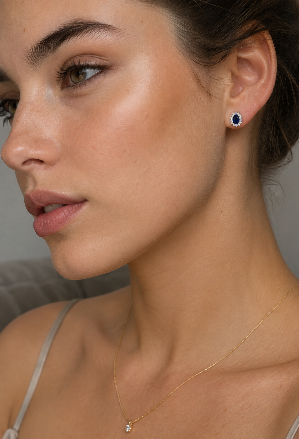 Close-up of a woman wearing delicate sapphire stud earring with a deep blue stone and a delicate gold chain necklace with a small clear stone, shown against a soft gray background.