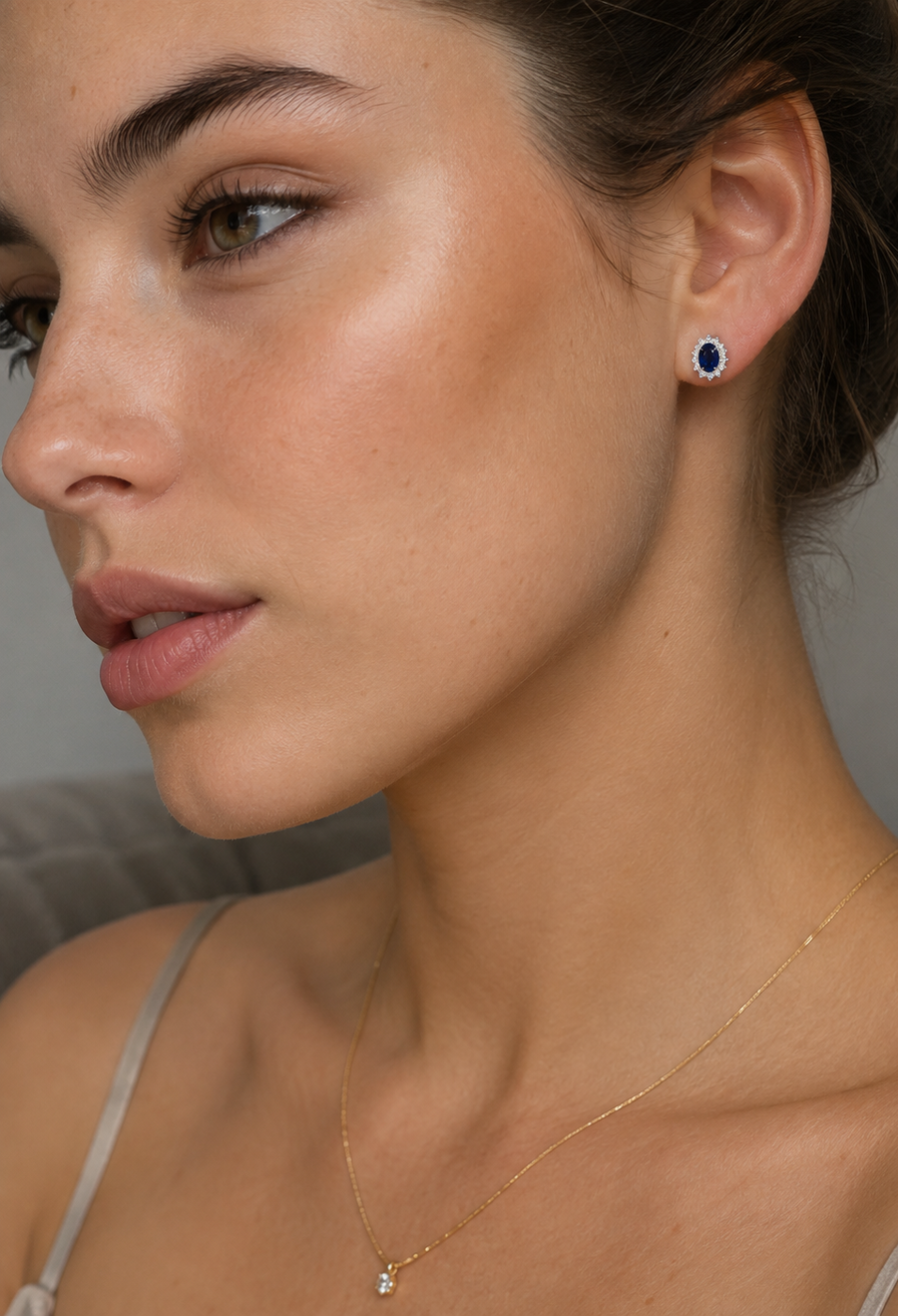 Close-up of a woman wearing delicate sapphire stud earring with a deep blue stone and a delicate gold chain necklace with a small clear stone, shown against a soft gray background.