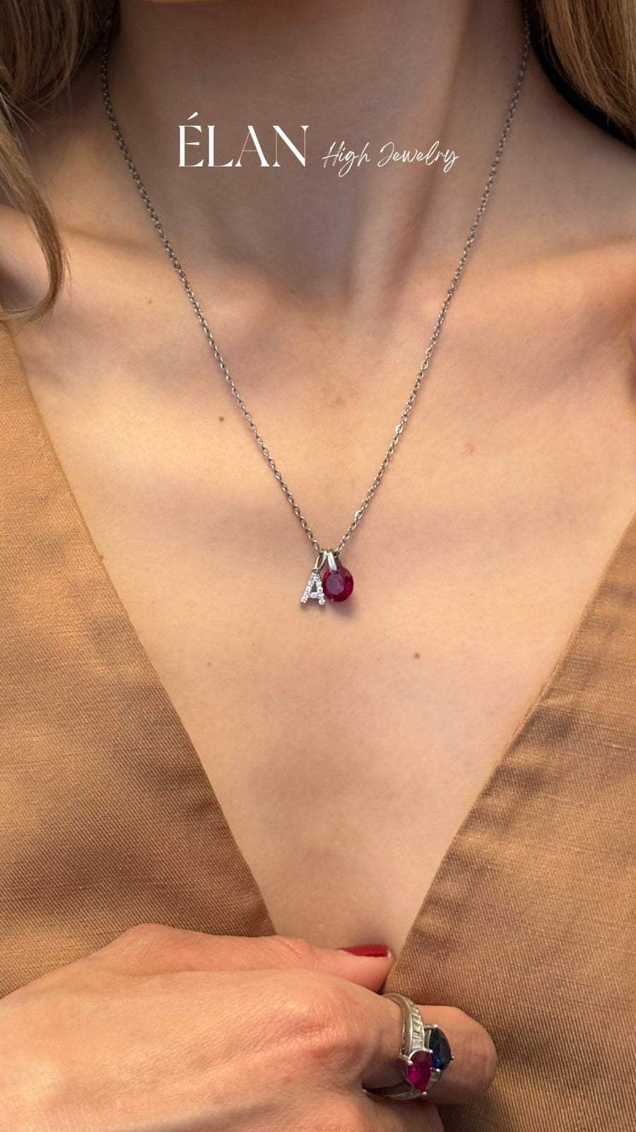 A close-up lifestyle shot of a model wearing a delicate silver necklace with a small letter 'A' pendant and a red gemstone, paired with a silver ring. The model is wearing a brown V-neck top, and the brand name 'ÉLAN High Jewelry' is visible at the top of the image.