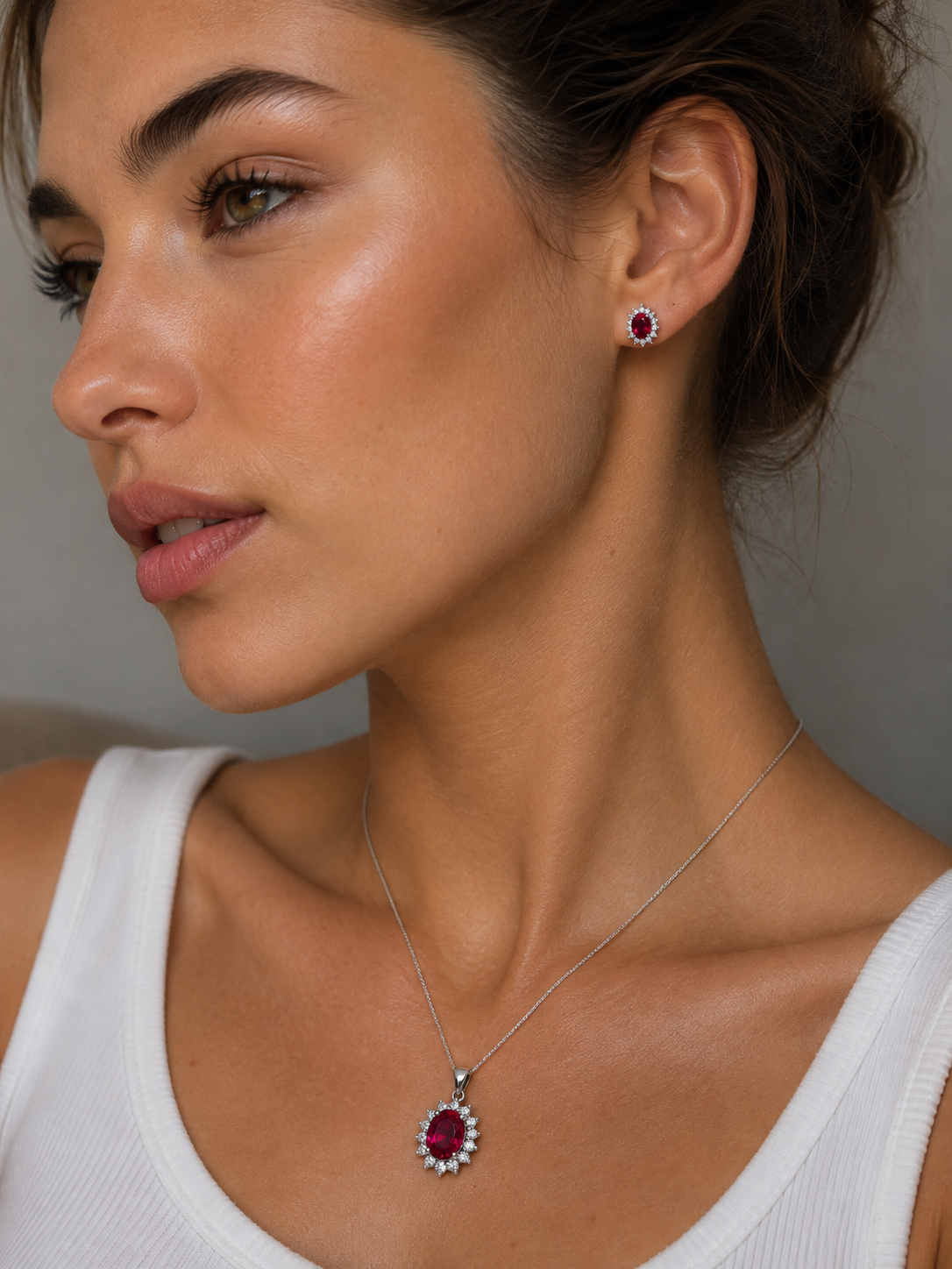 A close-up of a woman wearing a silver pendant necklace and matching stud earring, both featuring an oval cut red ruby gemstone surrounded by small clear diamonds in bulletproof setting Maison ÉLAN HIgh Jewelry
