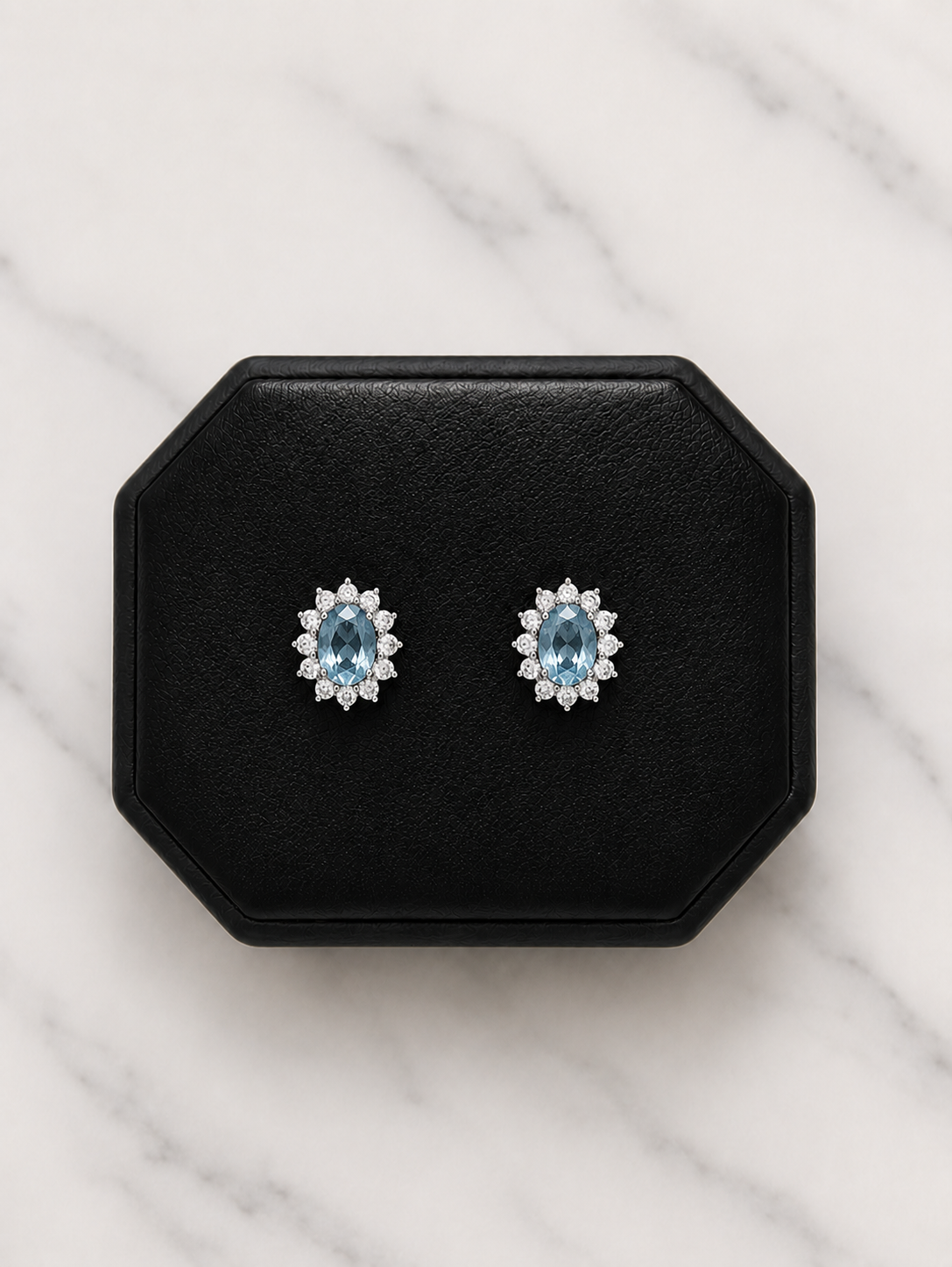 A pair of oval stud earrings with light blue center stones surrounded by small clear stones, displayed on a black octagonal jewelry box over a white marble surface.