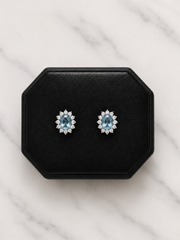 A pair of oval stud earrings with light blue center stones surrounded by small clear stones, displayed on a black octagonal jewelry box over a white marble surface.