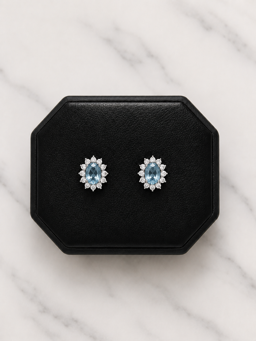 A pair of oval stud earrings with light blue center stones surrounded by small clear stones, displayed on a black octagonal jewelry box over a white marble surface.