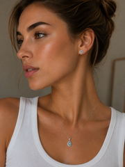 A close-up lifestyle shot of a woman wearing a delicate silver pendant necklace with a light blue oval stone and a matching stud earring. aquamarine