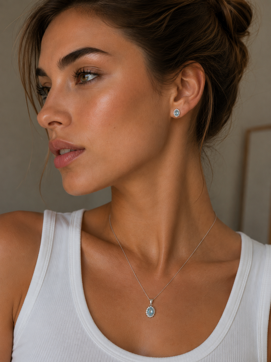 A close-up lifestyle shot of a woman wearing a delicate silver pendant necklace with a light blue oval stone and a matching stud earring. aquamarine