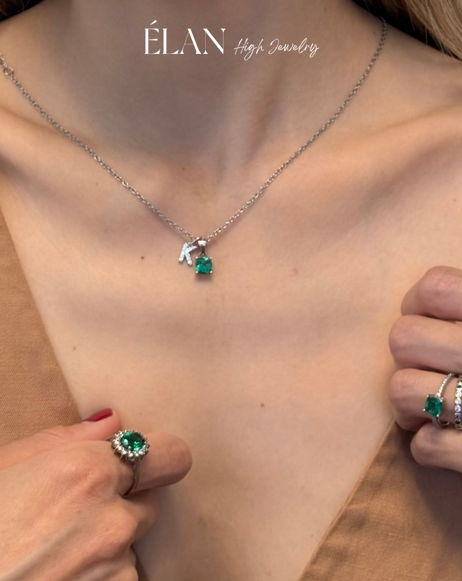 A close-up lifestyle shot of a model wearing a silver chain necklace with a crescent moon and green heart pendant, along with matching green gemstone rings. The brand name 'ELAN High Jewelry' is visible at the top.