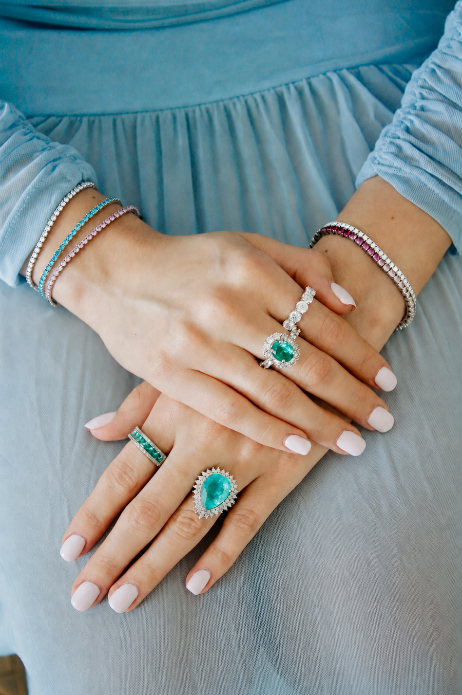 A close-up of a person's hands wearing multiple rings and bracelets, including a large emerald-cut green ring and smaller matching green stone jewelry, styled with a light blue dress. maison élan paraibas maison ÉLAN fine jewelry ring1